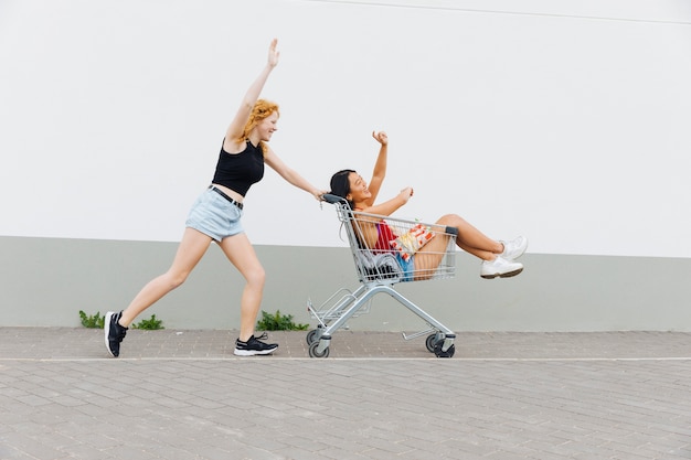 Woman rolling girlfriend with raised hands in shopping trolley 