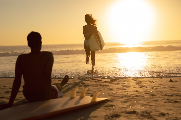 Woman Running with Surfboard while Man Relaxing on the Beach during Sunset