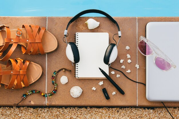 Woman's accessories composed on floor at pool, still life, view from above, summer fashion trend, vacation, headphones, notebook, sunglasses, sandals, seashell, pen, travel diary, bracelet, flowers