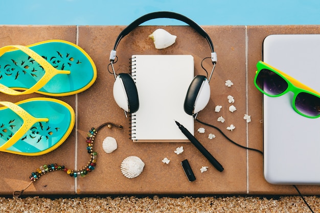 Woman's accessories composed on floor at pool, still life, view from above, summer fashion trend, vacation, headphones, notebook, sunglasses, sandals, seashell, pen, travel diary, bracelet, flowers