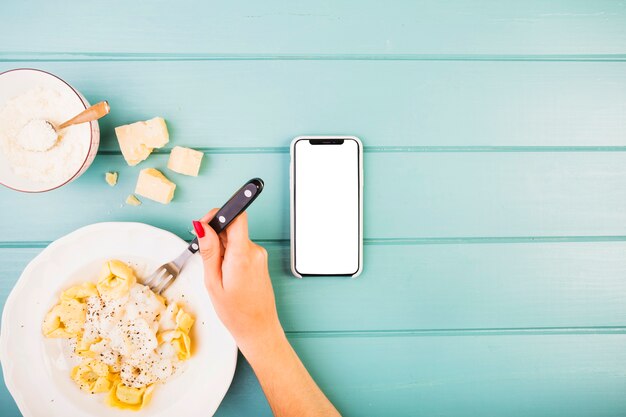 Woman's hand eating pasta with smartphone on desk