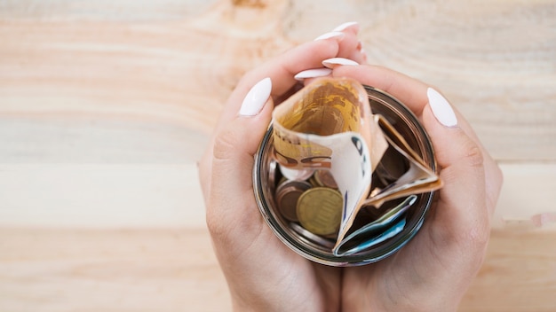 Woman's hand holding glass jar with euro notes and coins over wooden backdrop