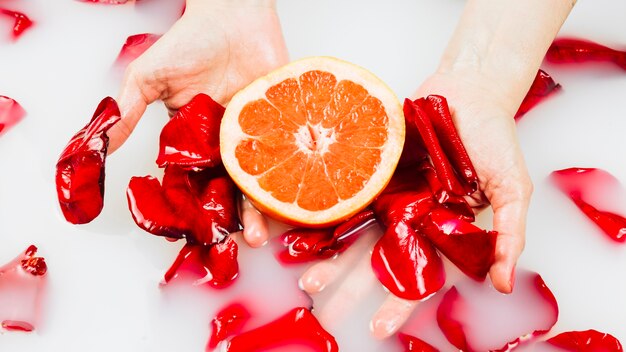 Woman's hand holding grapefruit and flower petals in spa bath with milk