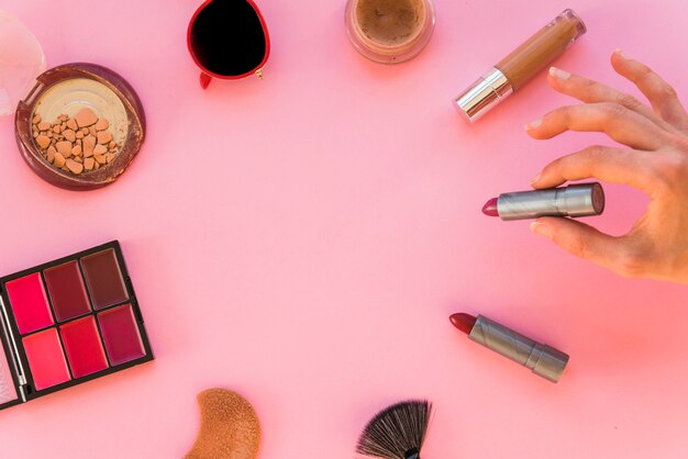 Woman's hand holding lipstick and various make up accessories on pink background