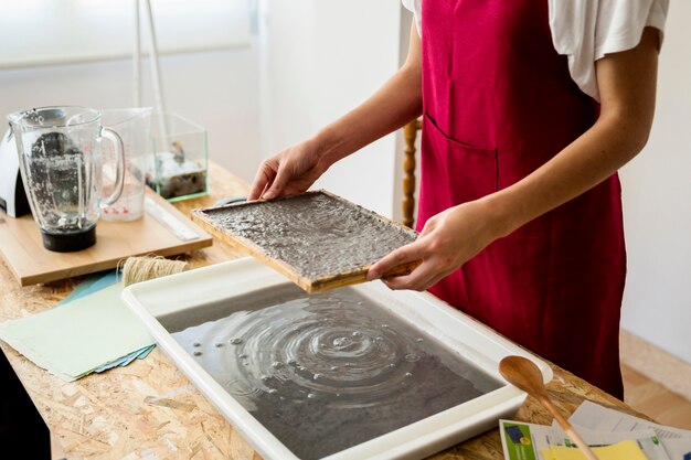 Woman's hand holding mold with paper pulp over tray in workshop