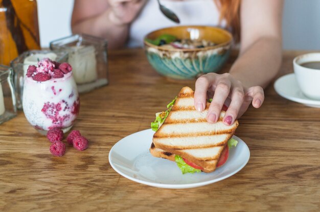 木製のテーブルに白いプレートからサンドイッチを持っている女性の手
