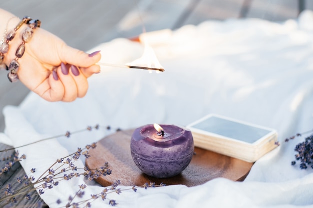 Женская рука в браслете с фиолетовыми длинными ногтями зажигает свечу со спичкой, рядом с колодой карт и цветами лаванды на белой и деревянной поверхности