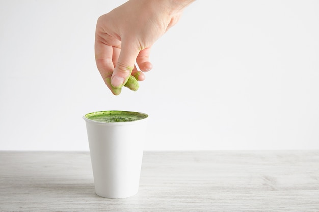 Woman's hand pours green organic premium matcha powder on top matcha latte in take away paper glass isolated on wooden table, white background