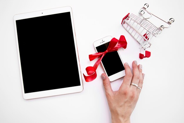 Woman's hand on smartphone near tablet and shopping trolley