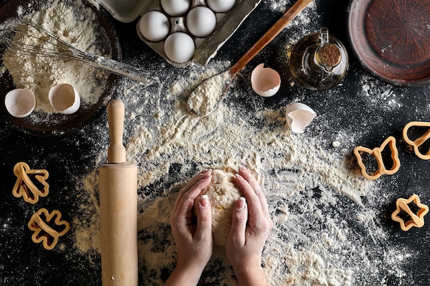 Woman's hands knead dough on table with flour, eggs and ingredients on black table. top view. still life. flat lay