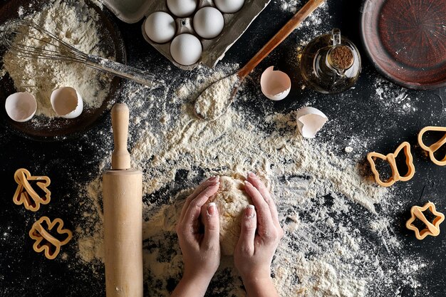 女性の手は、黒いテーブルの上に小麦粉、卵、材料を使ってテーブルの上で生地をこねます。上面図。静物。フラットレイ