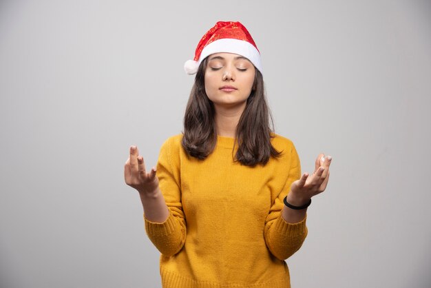 Woman in Santa hat doing meditation on gray wall. 