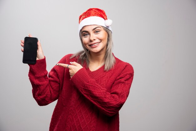 핸드폰을 가리키는 산타 모자에 있는 여자.