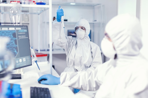 Woman scientist in coverall using micropipette in lab for coronavirus research. Team of microbiologists in research laboratory conducting experiment during global pandemic with covid19.