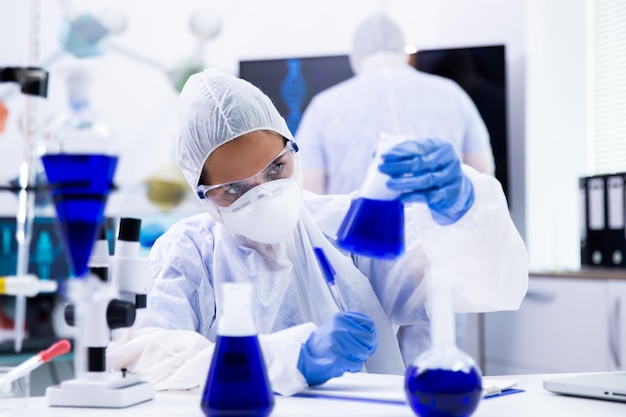 Женщина-ученый смотрит на стакан с синим веществом внутри. Коллеги по лаборатории, работающие сзади