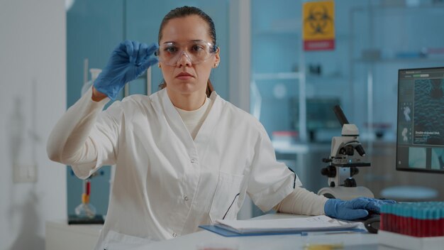 Woman scientist looking at blood sample on tray in laboratory, analyzing dna for diagnosis. Chemist with gloves and protective goggles holding glassware with substance for research.