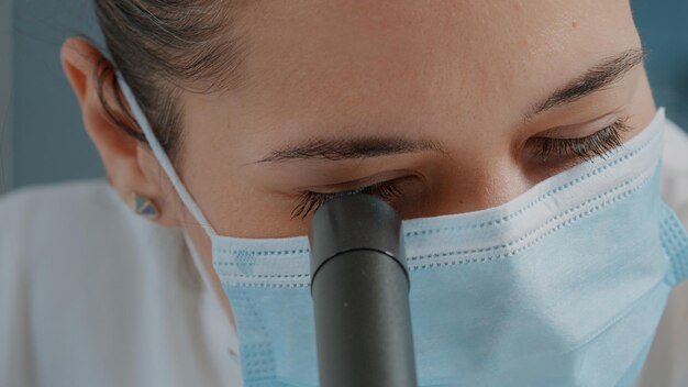 実験室での科学実験のために拡大鏡付きの顕微鏡を使用している女性科学者。フェイスマスクを着用し、科学ツールで顕微鏡レンズを使用するスペシャリスト。閉じる