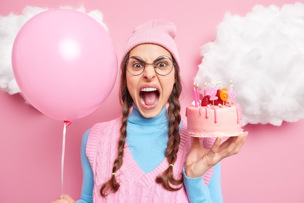 Free photo woman screams loudly keeps mouth widely opened being irritated because of something holds inflated balloon and birthday cake expresses negative emotions. celebration concept