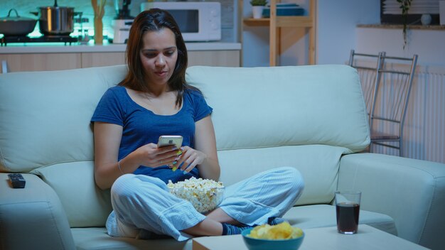 Woman scrolling on phone eating popcorn and watching a movie. Lonely amused happy lady reading, writing, searching, browsing on smartphone laughing amusing using technology internet relaxing at night.
