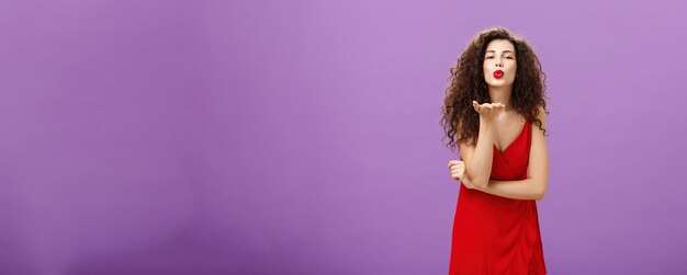 Woman sending passionate kiss being hopeless romantic standing in elegant red dress with curly hairs