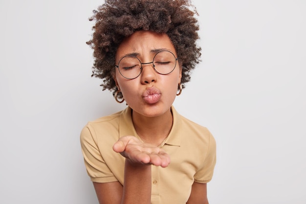 女性は空気のキスを送り、唇を丸く保ち、mwahは目を閉じ、丸い眼鏡をかけますベージュのTシャツは白で隔離されています。私の恋人になって。ボディーランゲージの概念。