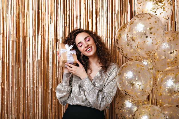 Woman in shiny blouse holding gift box with white bow on golden background