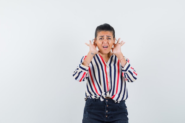 woman in shirt, skirt holding fingers behind ears and looking confused