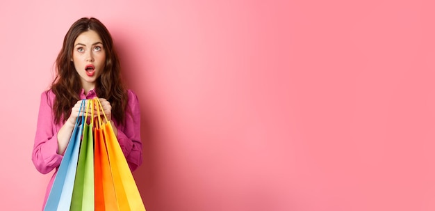 Free photo woman shocked of discounts looking up with amazed face holding shopping bags standing against pink b