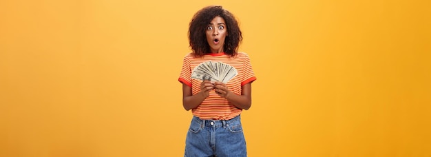 Free photo woman shocked finding lots of cash in safe portrait of surprised speechless goodlooking darkskinned female with curly haircut folding lips gasping holding money posing over orange background