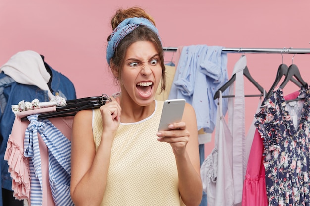 Donna shopping shoppaholic nel centro commerciale, con grucce di abiti eleganti, urla di rabbia e shock, utilizzando l'app di banking online su cellulare, sentendosi frustrato che il denaro non è disponibile nel conto bancario