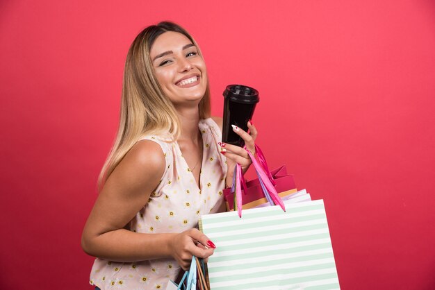 Woman showing her shopping bags and cup on red wall . 