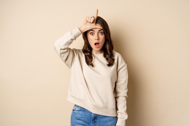 Woman showing loser sign on forehead, L word, standing over beige background