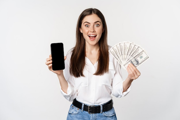Woman showing mobile phone screen and cash, money, concept of microcredit and bank loans, standing over white background.