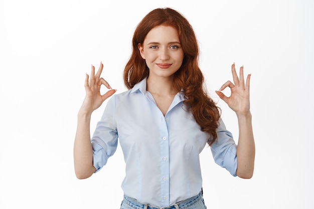 Бесплатное фото Женщина показывает жест ок с уверенным и напористым лицом, довольна хорошей вещью, хвалит выбор, стоит на белом