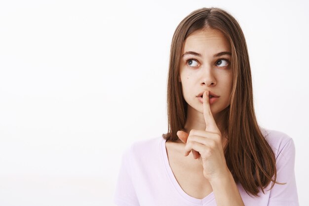 woman showing shush gesture while saying shh with index finger over mouth looking at upper left corner worried and intense