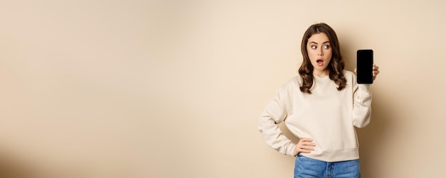 Woman showing smartphone screen and looking surprised at mobile phone standing in sweater over beige