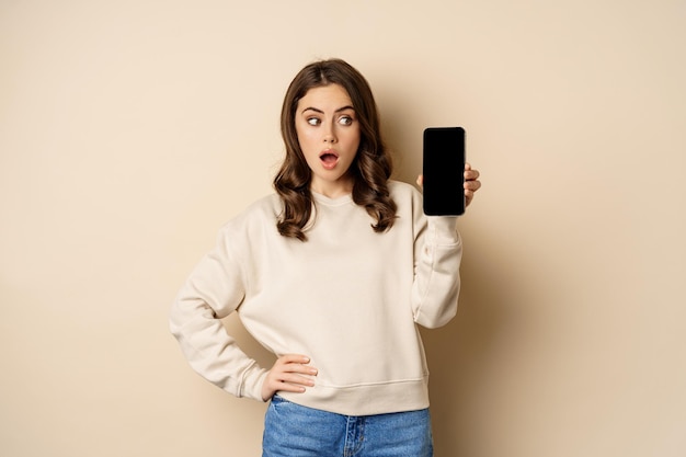 Woman showing smartphone screen and looking surprised at mobile phone standing in sweater over beige...