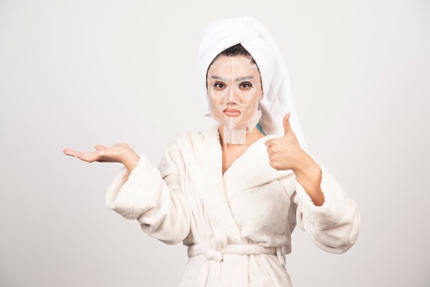 Woman showing thumb up and wearing bathrobe and towel with facemask.