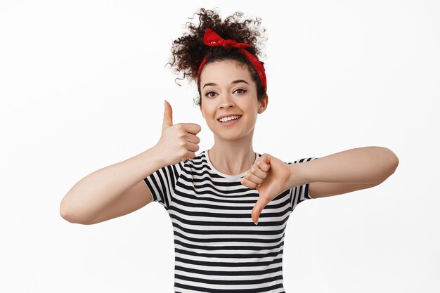 woman showing thumbs up and down, rating and feedback concept. standing in headband with combed curly hair and t-shirt on white