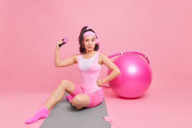 женщина показывает бицепс поднимает руку тренирует мышцы со спортивным оборудованием регулярно занимается спортом сидит на фитнес-коврике слушает музыку в наушниках использует швейцарский обруч с мячом
