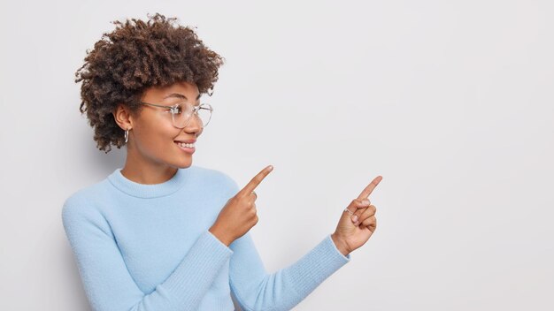 woman shows copy space demonstrates her choice wears spectacles and blue jumper isolated on white.