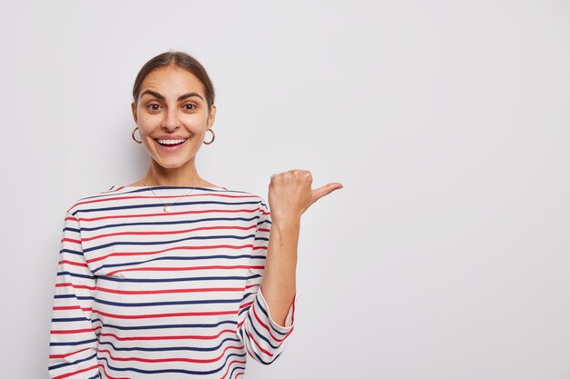 woman shows copy space demonstrates promotion points thumb away dressed in casual striped jumper on white