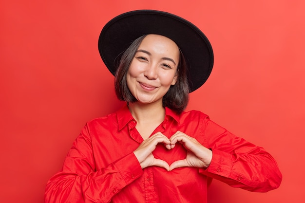 Free photo woman shows heart sign near chest i love you gesture wears black hat and shirt poses on vivid red