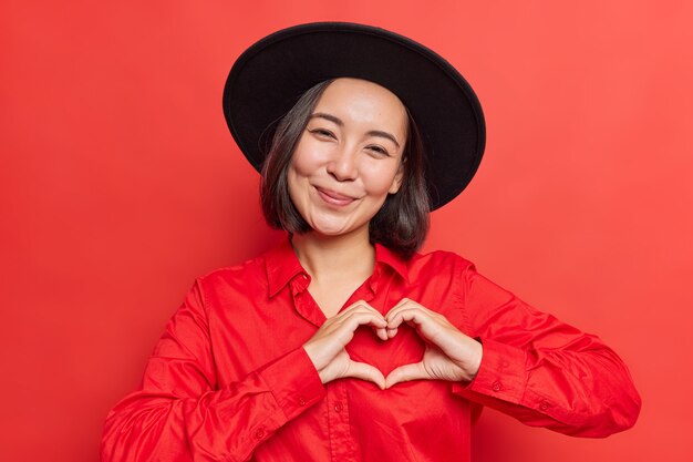  woman shows heart sign near chest I love you gesture wears black hat and shirt poses on vivid red 