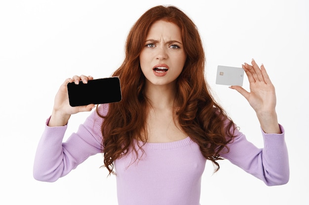 woman shows horizontal smartphone screen and credit card, frowning and looking disappointed, complaining about something on phone, standing on white