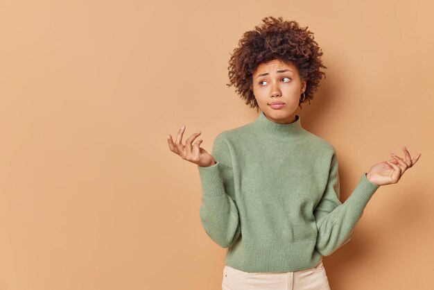 woman shrugs shoulders has indecisive expression cannot decide what to do has sad expression wears casual jumper isolated on beige with blank space for advertisement.