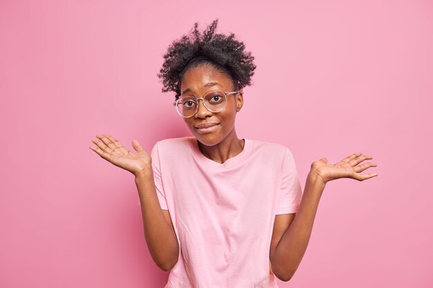 여자는 어깨를 으쓱하며 빈 손은 아무 말도 할 수 없다는 것을 보여줍니다. 실내는 분홍색 벽에 격리된 큰 광학 안경 캐주얼 티셔츠를 입고 있습니다