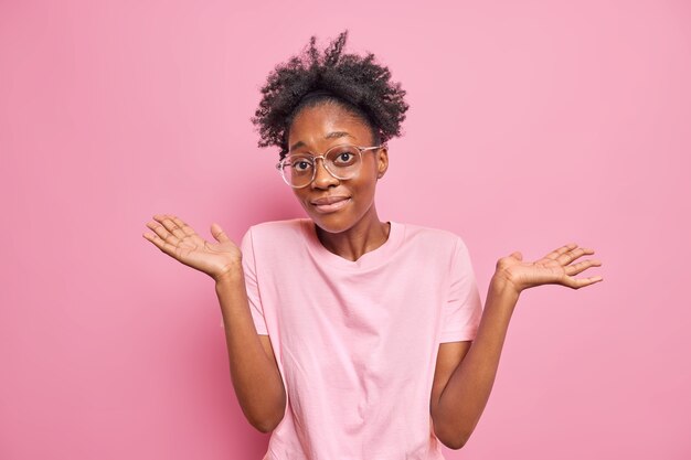 woman shrugs shoulders shows empty hands has no clue about something nothing to say stands unaware indoor wears big optical glasses casual t shirt isolated on pink wall