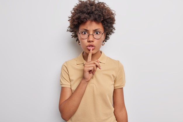 woman shushes with angry face makes taboo gesture tells to be quiet demands not to spread rumors wears round spectacles casual brown t shirt scolds person being too loud. Shut your mouth