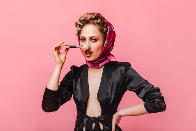 Женщина в шелковом халате и розовом шарфе держит кисть для макияжа и смотрит вперед